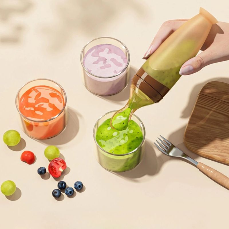 BabyHap™ Lepel-Knijpfles gebruikt om groene puree in een glazen pot te knijpen. Rondom de pot staan nog twee potten met oranje en paarse puree, en verschillende stukjes fruit liggen ernaast.