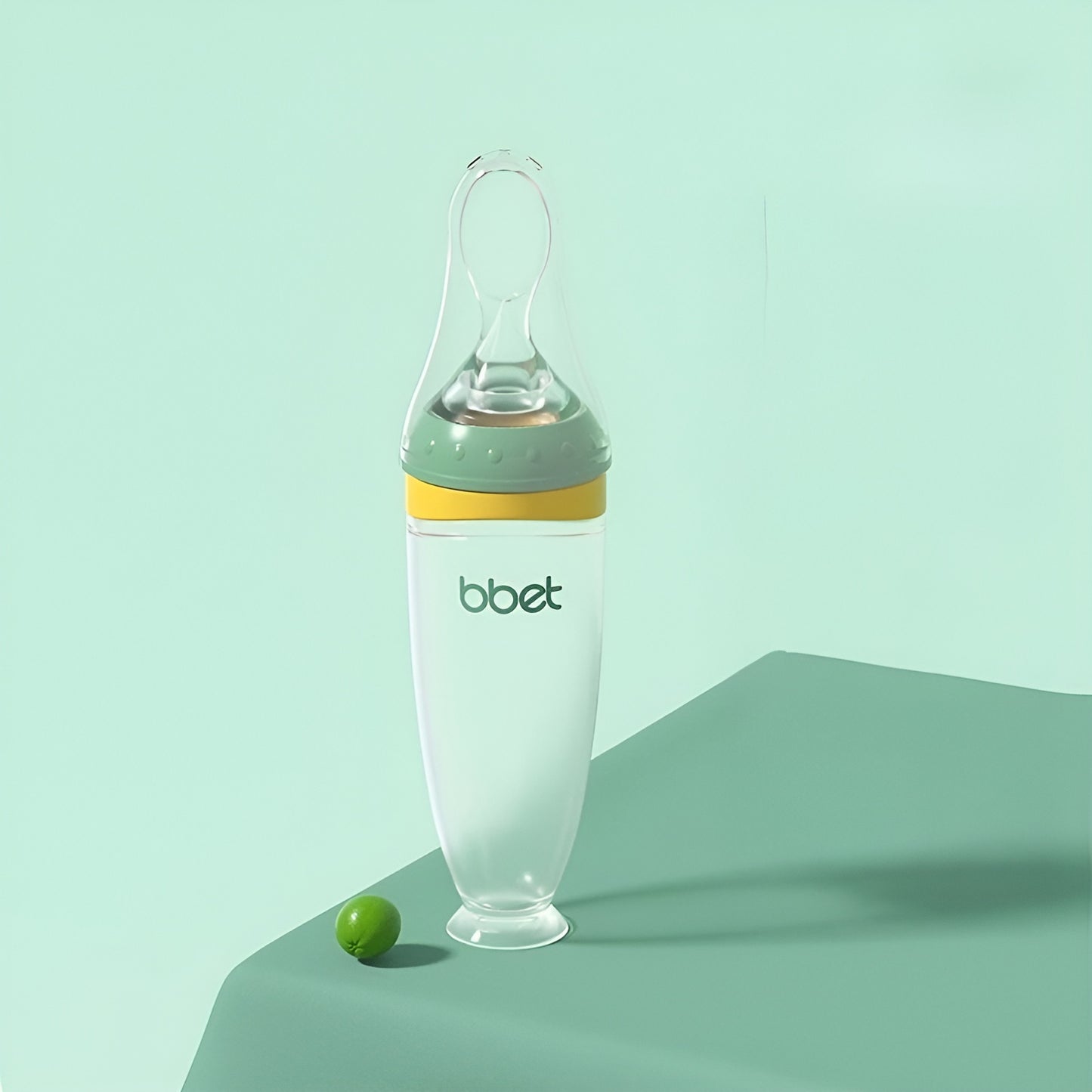 BabyHap™ Lepel-Knijpfles met groene en gele accenten op een groene ondergrond. De fles is ideaal voor stressvrij voeden, gemaakt van voedselveilige siliconen en eenvoudig schoon te maken.