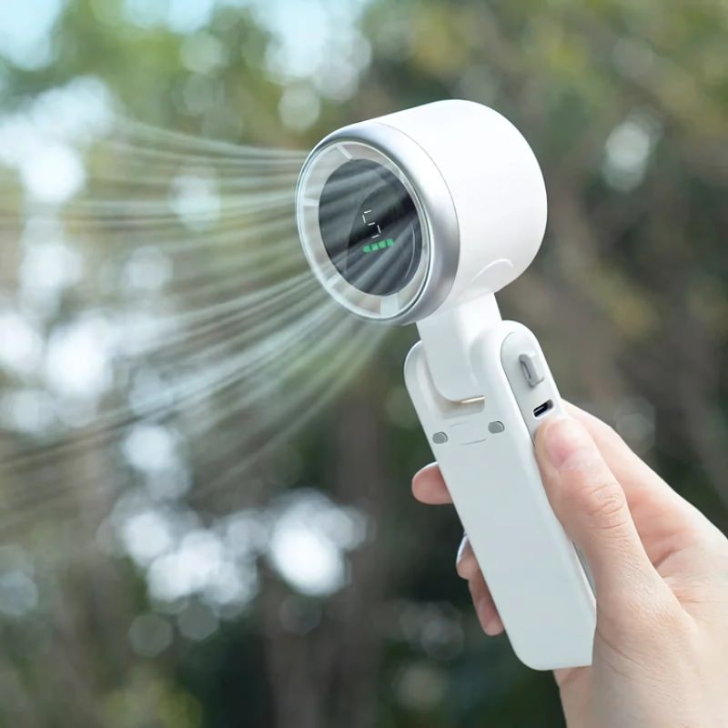Hand houdt een witte BreezeBuddy™ ventilator vast, blaast op volle kracht. Digitaal display toont stand 5. Compact formaat, zichtbaar Type-C oplaadpoort. Website: Inicala.nl.