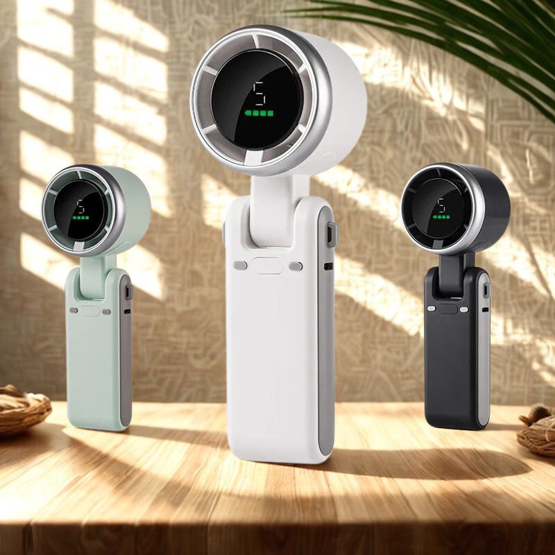 Drie BreezeBuddy™ ventilators op een houten tafel in zonlicht – wit, mintgroen en zwart model. Compact design met HD-display. Verkrijgbaar op Inicala.nl.