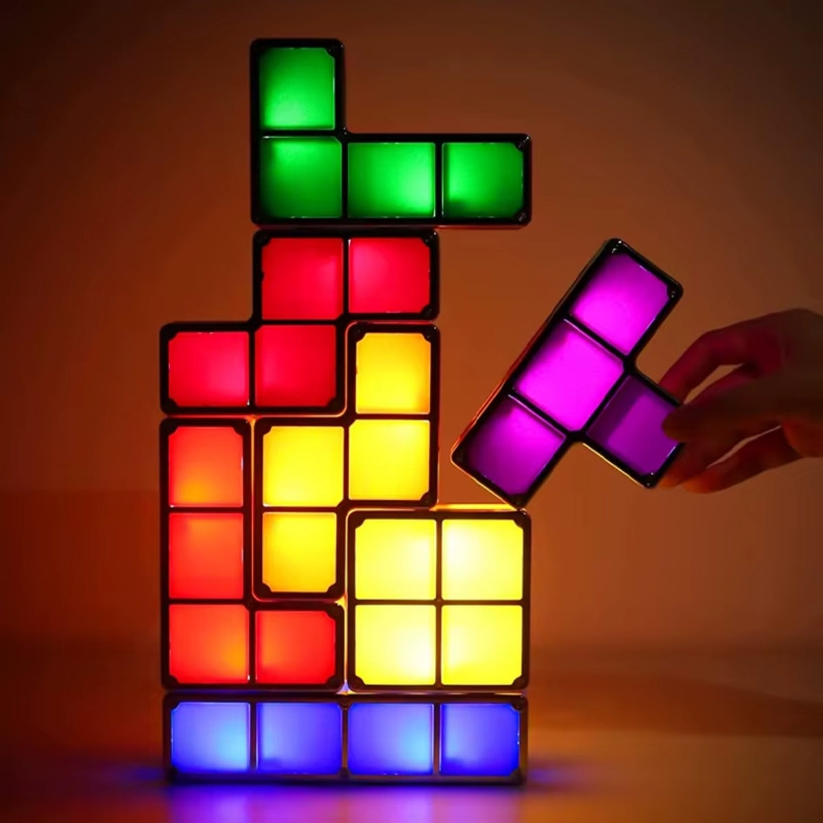 Een hand plaatst een paars TetraGlow™-blok op een stapel verlichte blokken in groen, rood, geel en blauw. De kleurrijke lampjes vormen een speels en creatief kunstwerk dat straalt in het donker. inicala