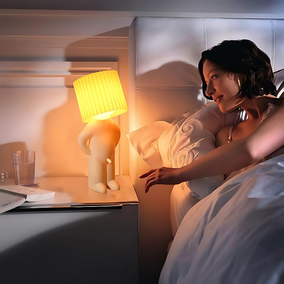 Een sfeervolle nachtlamp op een nachtkastje naast een vrouw die glimlacht. De Naughty Glow Guy™ verspreidt een warme gloed en voegt humor toe aan de slaapkamer. Een unieke combinatie van verlichting en fun. Ontdek meer op Inicala.nl!
