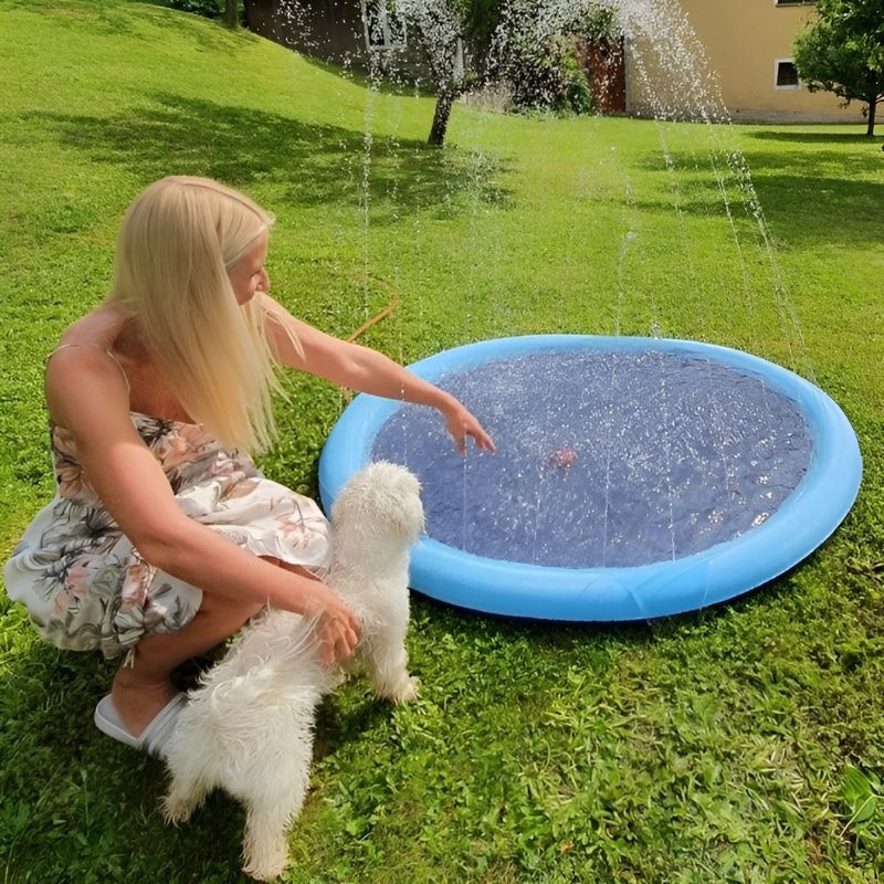 Een vrouw zit met haar witte hond naast een ronde, blauwe SplashPad™ FonteinMat. Waterstralen spuiten omhoog vanuit de rand, terwijl de hond nieuwsgierig naar het water kijkt. Ideaal voor verkoeling en speelmomenten in de tuin. inicala.nl