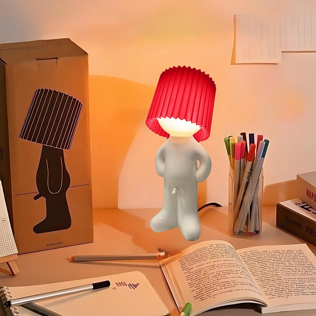 Naughty Glow Guy™ Grappig, brutaal, met een grenzeloze glimlach! Een speelse lamp met een opvallende rode kap, geplaatst op een bureau naast kleurrijke pennen en een open boek. Perfect voor humor en licht. Verkrijgbaar op Inicala.nl.