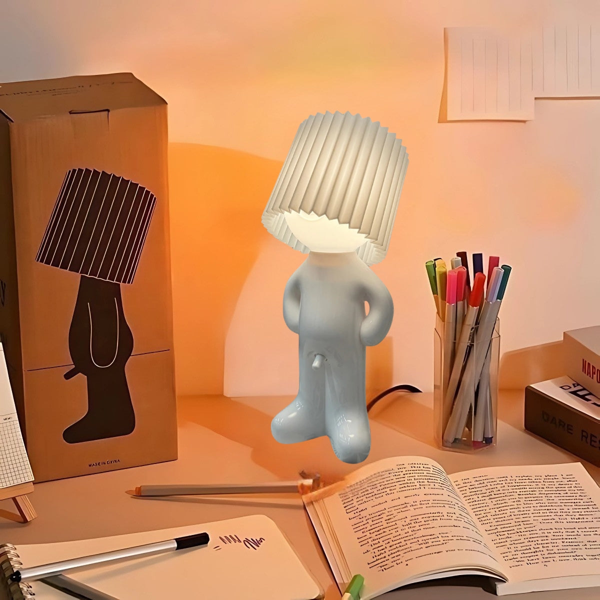 Naughty Glow Guy™ Grappig, brutaal, met een grenzeloze glimlach! Een speelse lamp met een strakke witte kap, omringd door een kleurrijk bureau en boeken. Voeg humor en stijl toe aan je interieur. Ontdek meer op Inicala.nl.