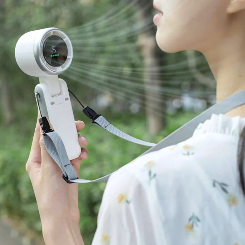 Vrouw gebruikt een kleine witte draagbare ventilator met nekkoord buiten. De luchtstroom is gericht op haar gezicht. Product BreezeBuddy™ – verkrijgbaar op Inicala.nl.