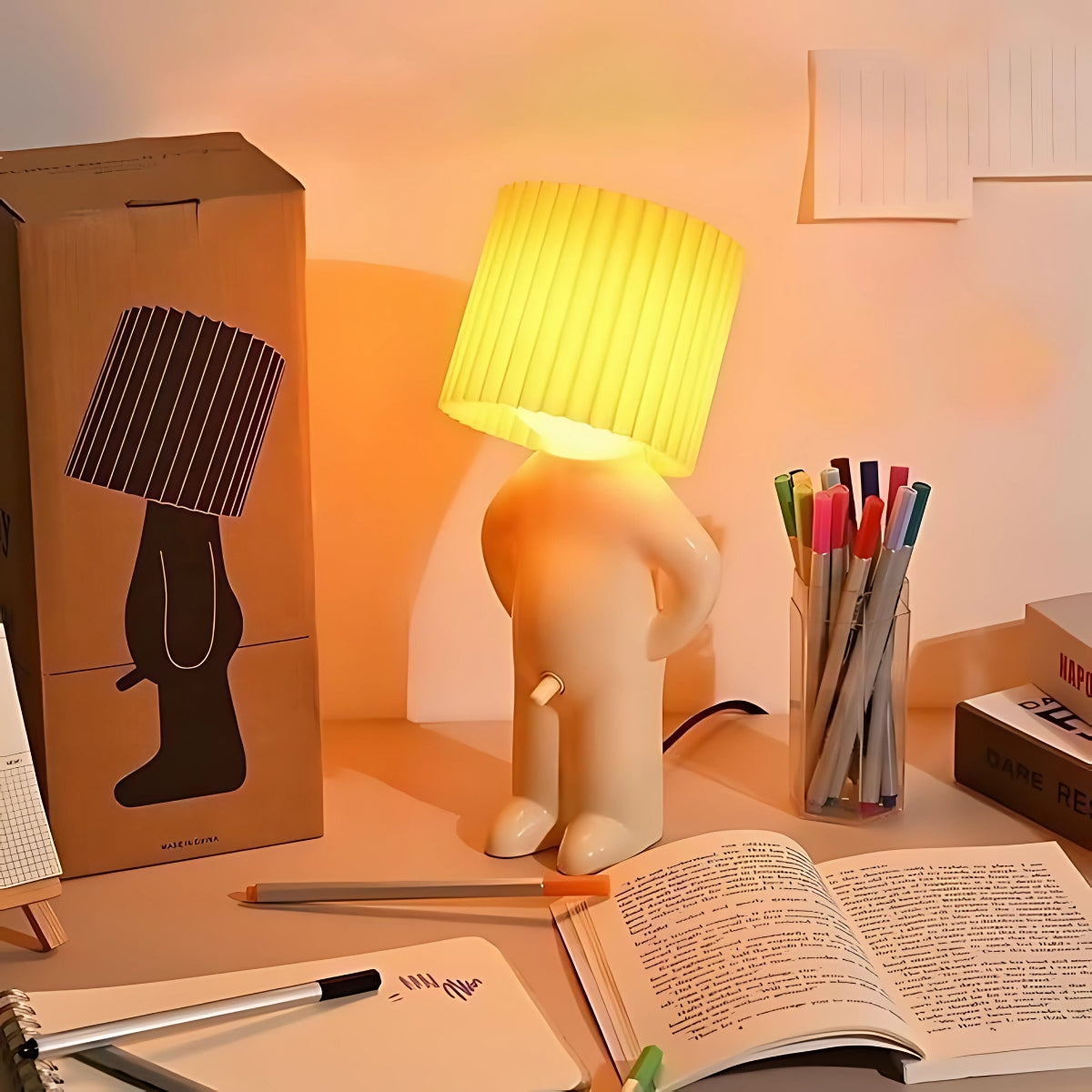 de Naughty Glow Guy™ op een bureau, omringd door kleurrijke pennen, boeken en notities. De lamp straalt een warme gloed uit en heeft een ondeugend ontwerp dat humor en sfeer toevoegt. Website: Inicala.nl.