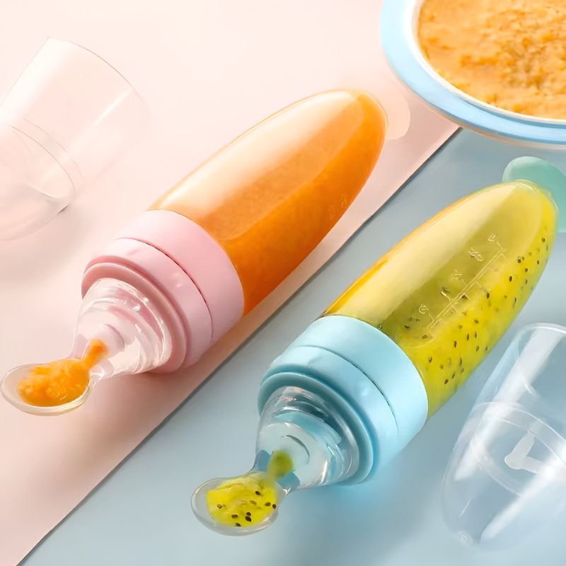 twee BabyHap™ Lepel-Knijpflessen gevuld met feloranje en groene puree. De flessen hebben zachte lepeltuiten en gekleurde doppen in roze en blauw. Een transparante beschermkap ligt ernaast.