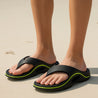 CloudRift™ – Ergonomische Slippers van Inicala.nl gedragen op het strand, met zwart design en neonlimoen accenten. Biedt comfort en grip tijdens warme dagen, perfect voor relaxte wandelingen of vakantie vibes.
