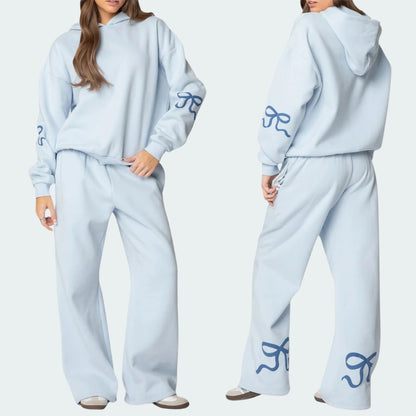 Luna Ribbon Tracksuit - Elegantie in Comfort Witte tracksuit met zachte blauwe strikaccenten op mouwen en broekspijpen. Ideaal voor een frisse, casual look. Shop exclusief o