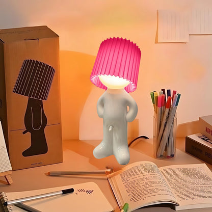 Naughty Glow Guy™ | Grappig, brutaal, met een grenzeloze glimlach! Een speelse lamp met een opvallende roze kap, geplaatst op een bureau naast kleurrijke pennen en een open boek. Perfect voor humor en sfeer. Ontdek meer op Inicala.nl.
