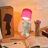 Naughty Glow Guy™ | Grappig, brutaal, met een grenzeloze glimlach! Een speelse lamp met een opvallende roze kap, geplaatst op een bureau naast kleurrijke pennen en een open boek. Perfect voor humor en sfeer. Ontdek meer op Inicala.nl.