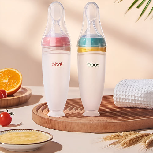 Twee BabyHap™ Lepel-Knijpflessen, één met roze en één met groene schroefdop, staan rechtop op een houten bord, omringd door een sinaasappel, tomaten en een witte handdoek, klaar voor gebruik.